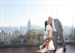 Lovely couple from the UK kissing on Top of the Rock with the Empire State building behind them. She has a beautiful white dress and is holding a bridal bout of red flowers.